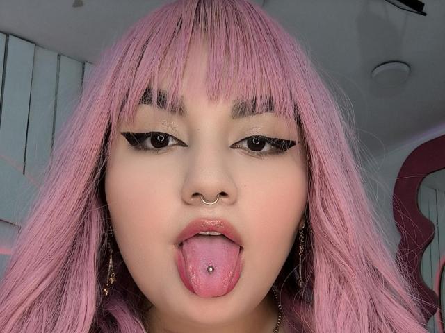 MarlaBriard has pink hair, black eyeliner, a nose ring, and her tongue out with a tongue piercing. Her pink lips are slightly parted. She's looking at the camera.