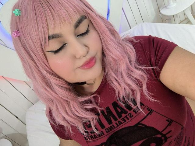 MarlaBriard with pink hair, eyeliner, nose ring, and pink lips, wearing a maroon t-shirt, sitting on a white bed, looking down, with cute hairpins.