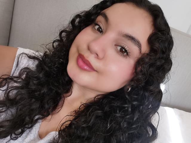 TaniaRichmond has curly black hair, brown eyes, and pink lipstick, wearing a grey shirt, with a soft smile, looking slightly to the right. Her boobs are not visible.