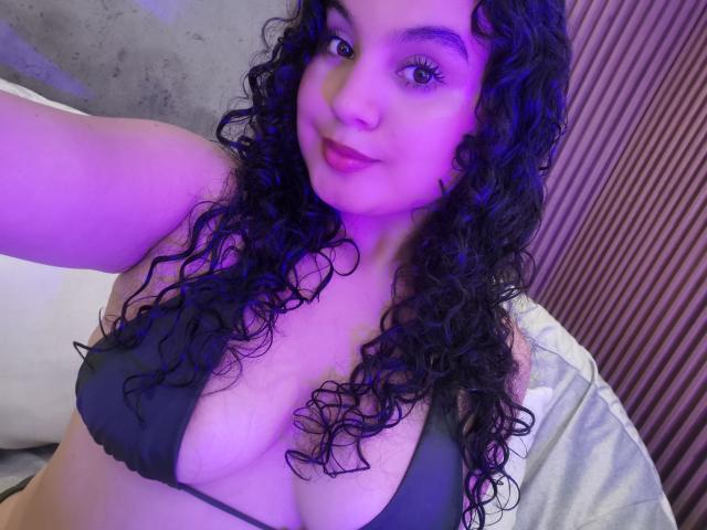 TaniaRichmond with curly black hair, purple lighting, and a black bikini top showing her big boobs, smiling slightly at the camera. She has a light skin tone and full lips.