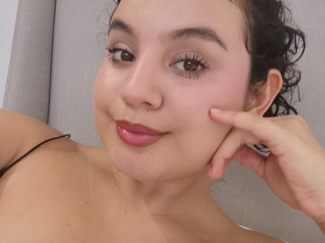 TaniaRichmond has curly black hair, pink cheeks, and is smiling with pink lipstick. She's touching her cheek with her right hand, showing black eyeliner and long eyelashes.