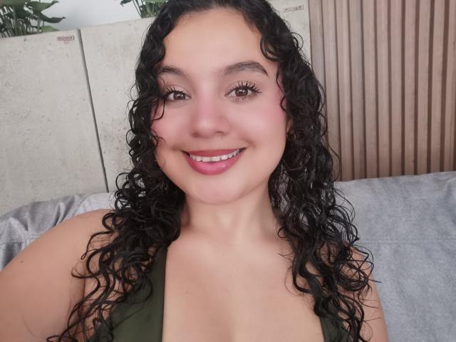 TaniaRichmond has curly black hair, brown eyes, pink lipstick, and is smiling at the camera with her boobs slightly visible in a green top.