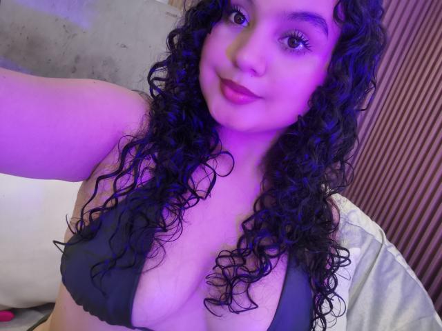 TaniaRichmond with curly black hair, purple-lit face, and small black bikini top showing her tits, smiling at the camera. She has pink lipstick and a cute, sexy look.
