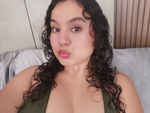 TaniaRichmond with curly black hair, pouty pink lips, and green top, showing her tits in a selfie. She has a light skin tone and brown eyes.