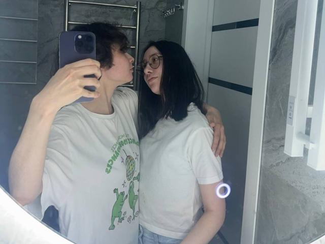 TheaRollock, com cabelo preto na altura dos ombros, óculos e camiseta branca, está ao lado de TheaRollock, com cabelo castanho encaracolado, camiseta branca e a mão no quadril, em uma selfie no espelho do banheiro..