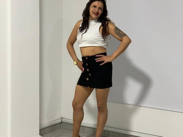 AriannaSeete with long dark hair, white sleeveless crop top, black button-up skirt, tattoo on left arm, smiling, hands on hips, wearing a yellow watch, standing barefoot.