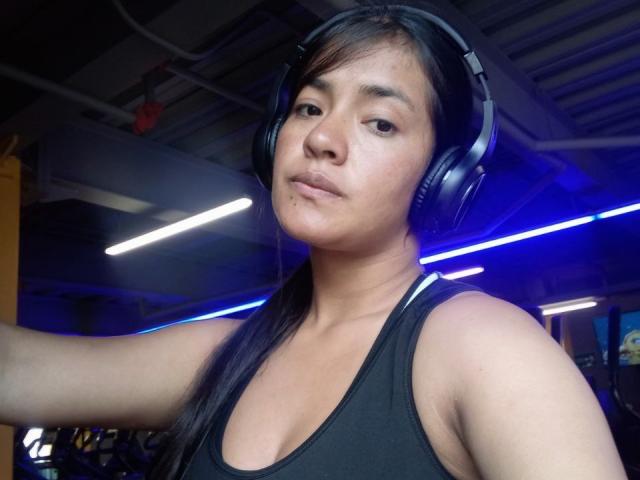 ArianithaXox with black headphones, medium tits, black tank top, long black hair, slightly pouty face, in a gym with blue lights.