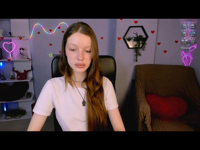 Khloeee with long brown hair, white tee, and dog tag necklace, sitting in chair, looking down with pouty lips, neon heart and deer wall decor.