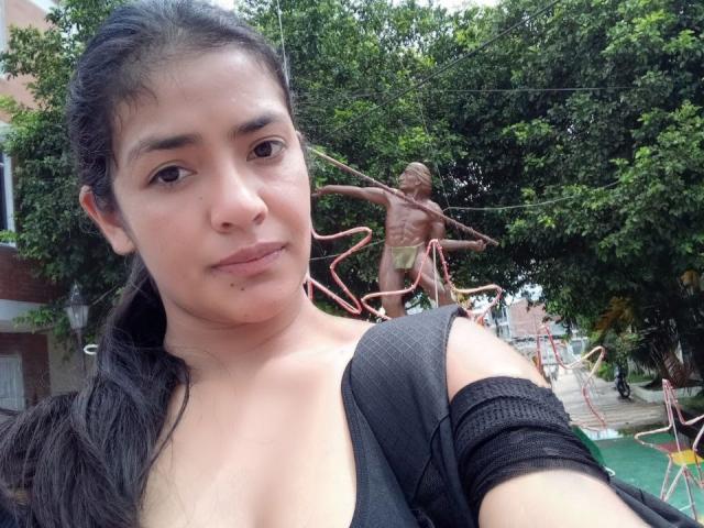 ArianithaXox with dark hair, light skin, and small tits, wearing a black top, stands in front of a nude, spider-like statue with long arms in a green outdoor space.