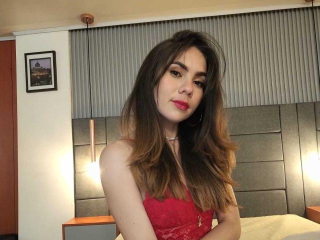LindaPerez with long brown hair, red lace dress, pink lipstick, hoop earrings, and a subtle smile, sitting in a hotel room with a grey headboard and hanging lights.