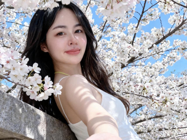 XweiWei, met lang zwart haar, een witte tanktop en een zachte glimlach, leunt tegen een rots, omringd door roze kersenbloesems, met een blauwe lucht op de achtergrond, en kijkt omhoog naar de camera..