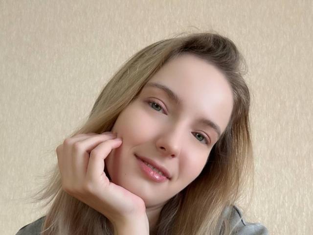 Nicole Spritzer, con i capelli castano chiaro, gli occhi verdi e un leggero sorriso, appoggia la guancia sulla mano, mostrando un'espressione carina e rilassata. Le sue labbra rosa sono leggermente dischiuse..