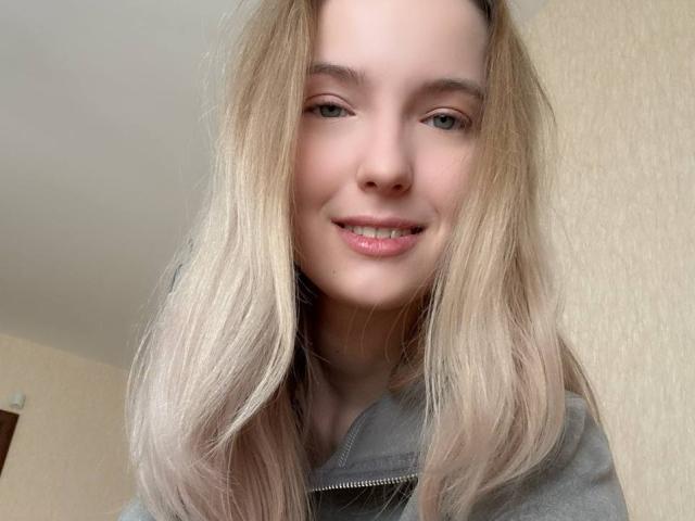 NicoleSpritzer has blonde hair, light skin, green eyes, and is smiling with slightly parted lips. She's wearing a grey hoodie, and her tits are not visible.
