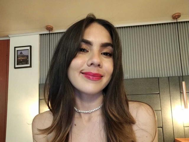 LindaPerez has long brown hair, red lipstick, and a pearl necklace, smiling at the camera with her tits barely visible, wearing a strapless top in a warm-lit room.