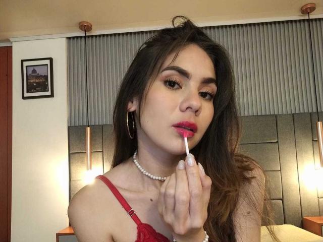 LindaPerez with long brown hair, red lipstick, and red bra, applying pink lipstick with a finger. She's wearing a pearl necklace and large hoop earrings.