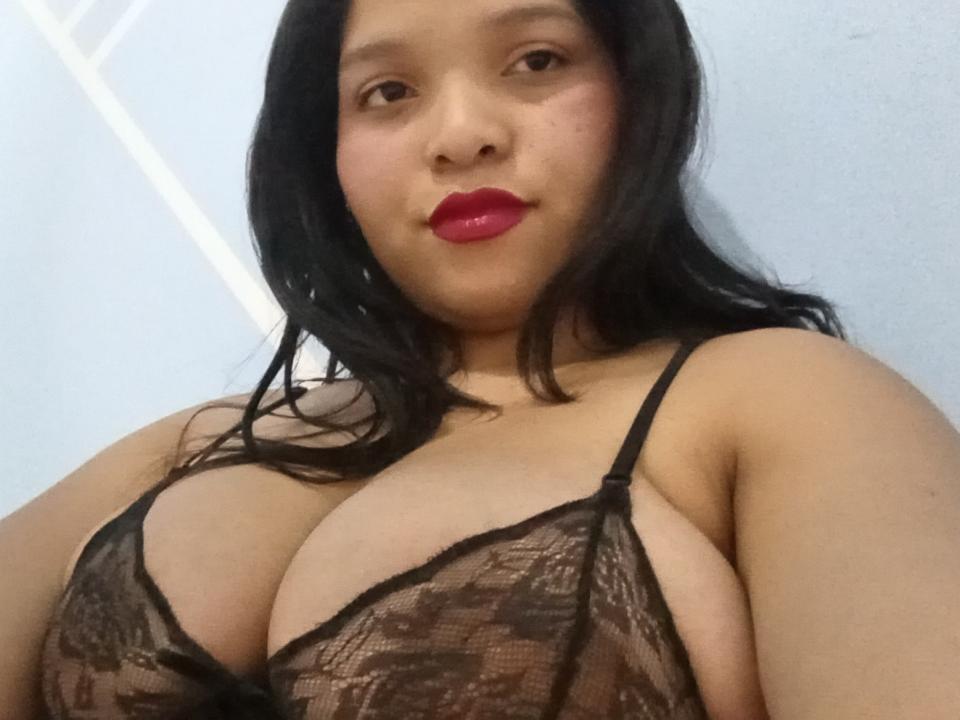 LunaSweett with red lips, black lace bra, large tits, and long black hair, looking at the camera. Her skin is light brown, and she's smiling slightly.