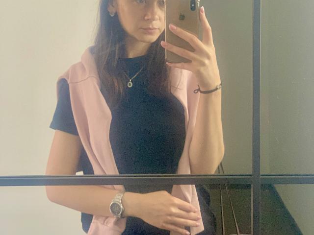 Dalaila with medium tits, wearing a black shirt and pink vest, taking a mirror selfie with a gold iPhone. She has a watch, necklace, and bracelet.