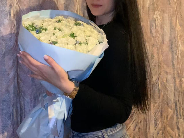Dalaila, con lunghi capelli neri, una camicia nera a maniche lunghe e jeans blu, tiene un bouquet di fiori bianchi con foglie verdi, indossa un orologio d'oro, sullo sfondo di un muro strutturato..