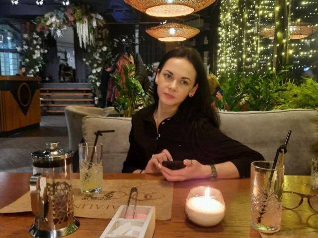 Dalaila, black-haired, sitting at a wooden table with a candle, glass, and drink, wearing a black shirt, looking at her phone, in a cozy, dimly lit cafe.