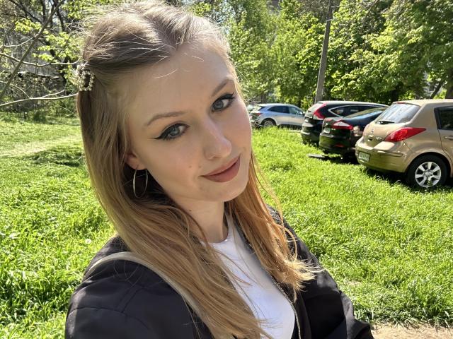 Nikollia has blonde hair, blue eyes, and a slight smile. She's wearing a black jacket over a white shirt, with small hoop earrings. Her tits are partially visible.