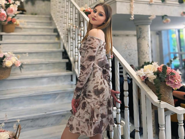Nikollia, con lunghi capelli castani, indossa un abito floreale con le spalle scoperte, è in piedi su una scala bianca, guarda indietro con il rossetto rosa e un sorriso appena accennato, con le mani vicino ai glutei..
