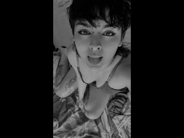 LilithDemmon with short dark hair, nose ring, and tattoos, wearing a lace bra, looking up with tongue out, kneeling on a bed, black-and-white photo.