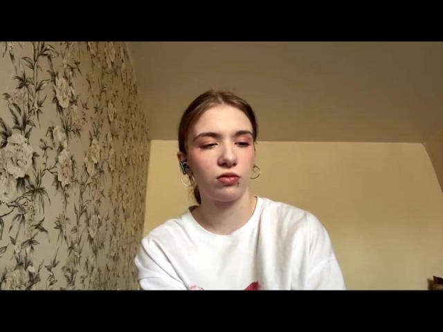 EviaMarcot with pouty lips, light makeup, and pink eyeshadow, wearing a white shirt, small hoop earrings, and brown hair pulled back. Background is floral-patterned wallpaper.