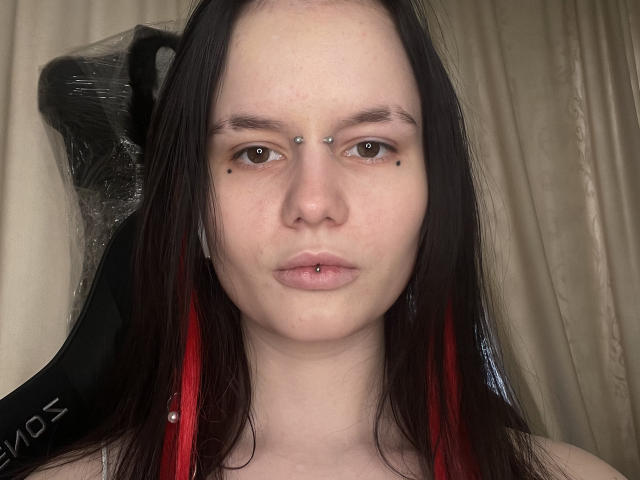 KaykaSunder with long black and red-dyed hair, piercing her nose and lower lip, has black moles on her upper lip and two small piercings above her brown eyes.