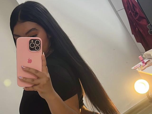 AlisBella with long black hair, holding pink iPhone, taking mirror selfie, showing black top, nails painted pink, holding phone with left hand, right arm covering lower part of face.