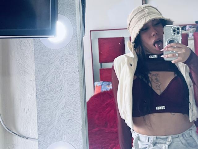 GeraldineSuan with tongue out, wearing a beige hat, open white vest, maroon bra, and light jeans, showing tits with tattoos above, taking a mirror selfie.