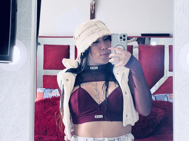 GeraldineSuan with dark hair, wearing a beige bucket hat, white vest, and maroon bralette, showing her tits, holding a phone, red pillows in the background.