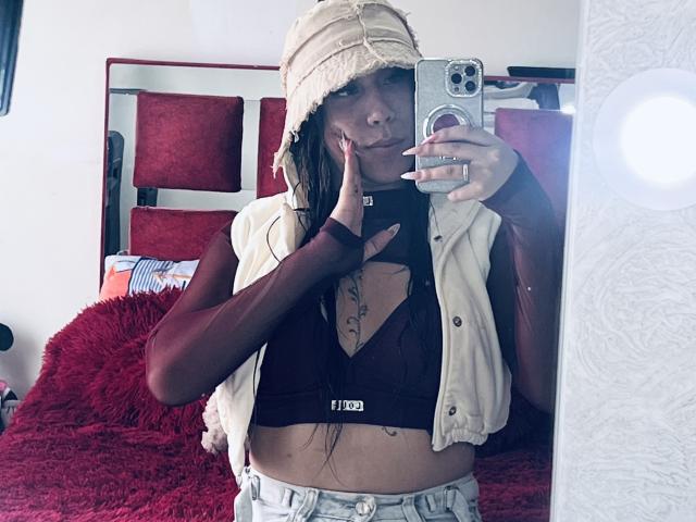 GeraldineSuan with long brown hair, wearing a beige bucket hat, maroon long-sleeve top, black bra, white vest, and blue jeans, holding a phone, showing off her tits.