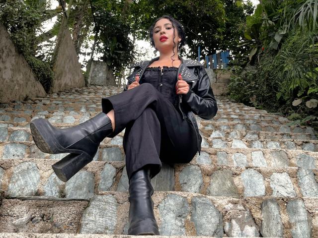 LilithStygian sits on stone steps, wearing black leather boots, a studded jacket, and black dress, legs crossed, hands on jacket, red lipstick, black hair, green eyes, looking down.