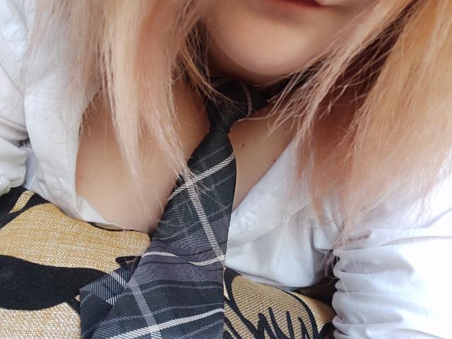 MinistraX with blonde hair, white shirt, and black plaid tie, showing cleavage, her tits slightly visible under the shirt. Her face is partially blurred. She's sitting on a patterned couch.
