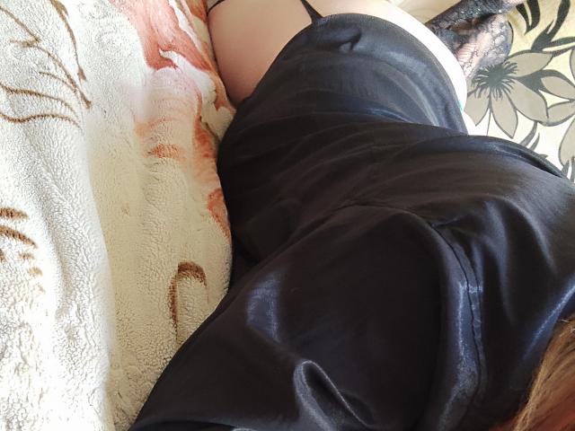 Les fesses de MinistraX sont partiellement couvertes par une robe en satin noir, et elle est allongée sur une couverture blanche à motifs floraux, le dos tourné vers la caméra. Son soutien-gorge en dentelle noire est visible..