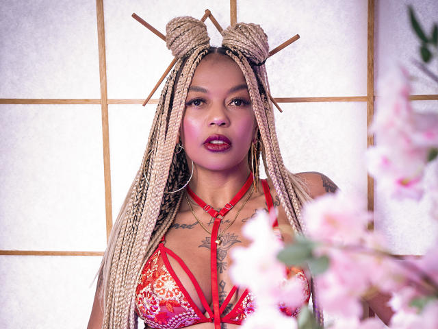 KenyaCarper with long blonde braids, red lace-up top, visible tattoos, red lipstick, pink blush, wooden hair sticks, pink cherry blossoms in foreground, standing in front of white panel.