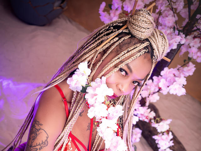 KenyaCarper with blonde braids, red bra, and cherry blossoms in her face, has a tattoo on her arm and a bun on top of her head, smiling at the camera.