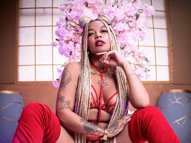 KenyaCarper with long blonde braids, red lingerie, tattoos, and red thigh-high boots, sitting with legs spread, hand on chin, pink cherry blossom headpiece, looking at camera.