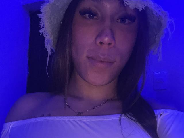 GeraldineSuan with blue-lit face, wearing a white off-shoulder top and a fuzzy white hat, has long brown hair, a nose ring, and a necklace with a small pendant.