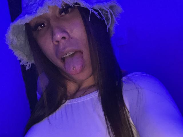 GeraldineSuan with long brown hair, tongue out, white top, and white frayed bucket hat, blue-lit face, visible tongue piercing, and a necklace, looking into the camera.