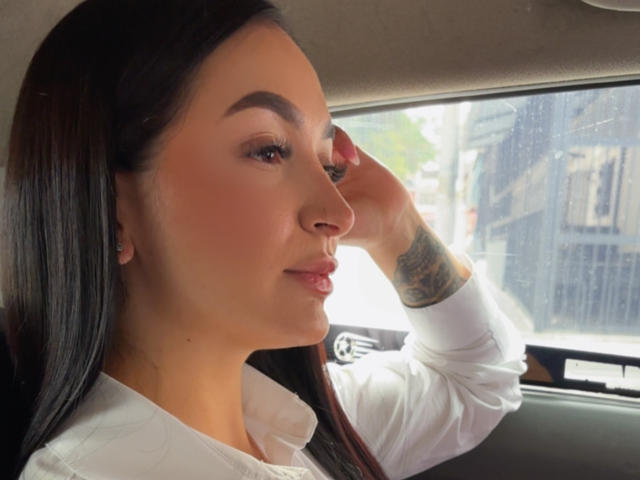 PamelaLiss with long black hair, wearing a white shirt, has her right hand up, showing a tattoo on her wrist, looking out of a car window with a slight smile.