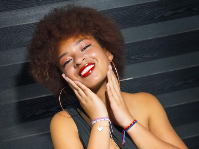 NiaZuri mit rotem Lippenstift, lächelnd, mit den Händen auf den Wangen, trägt Ohrringe im Stecker-Stil, ein violettes Armband und ein Herz-Armband, vor einem dunklen Holz Hintergrund, wirkt sie glücklich und selbstbewusst..