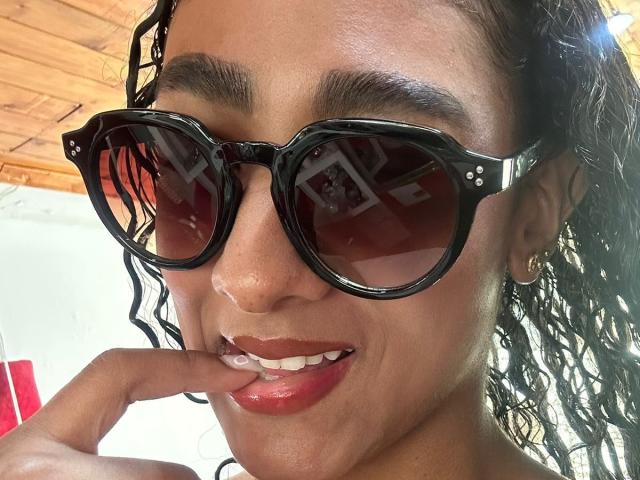 NatalyBlonder with dark curly hair, wearing black sunglasses, touching her glossy red lips with her finger, sporting a small gold earring, looking sexy and confident.