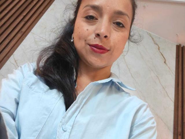 StangelarRivera, avec une peau brun clair, des cheveux noirs et ondulés attachés en queue de cheval, portant une chemise bleu clair à boutons, du rouge à lèvres, de petites boucles d'oreilles, et souriant légèrement..