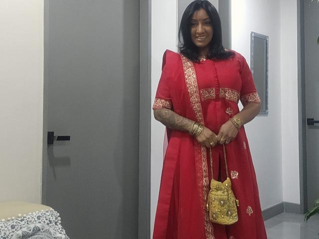 Nahirr with medium brown skin, black hair, wearing a red sari with gold trim, holding a yellow bag with gold studs, smiling, wearing gold bangles.