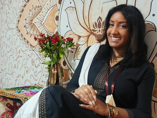 Nahirr, smiling, with dark hair, wearing a black traditional outfit, white scarf, gold necklace, and bracelet, holding hands, sitting with red flowers and colorful fabric on the left.