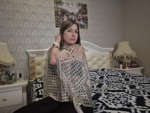 LoriEla with brown hair, wearing a sheer lace shawl, black pants, sitting on a bed with a black and white patterned blanket, white headboard, and two lamps.