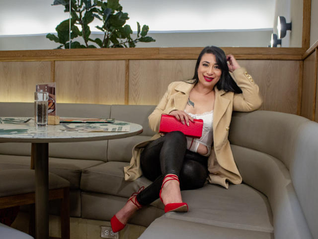KarimeLeeX with black hair, red lipstick, tan coat, white top, black leather pants, red heels, holding pink clutch, sitting on gray booth, smiling, tattoo on right chest.