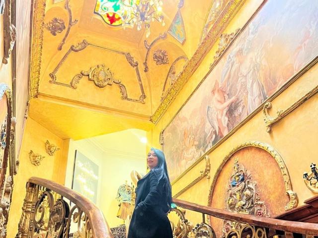 KarimeLeeX stands on ornate staircase, wearing black hijab and long-sleeve top, looking up at colorful stained glass ceiling, surrounded by gold-accented walls and large, detailed paintings.