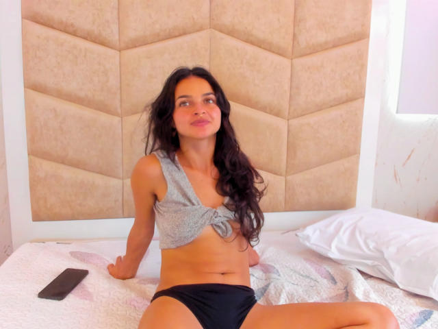 LucyySky with long black hair, wearing a tied gray top and black panties, sitting on a bed with a white pillow and black remote, looking to the side.